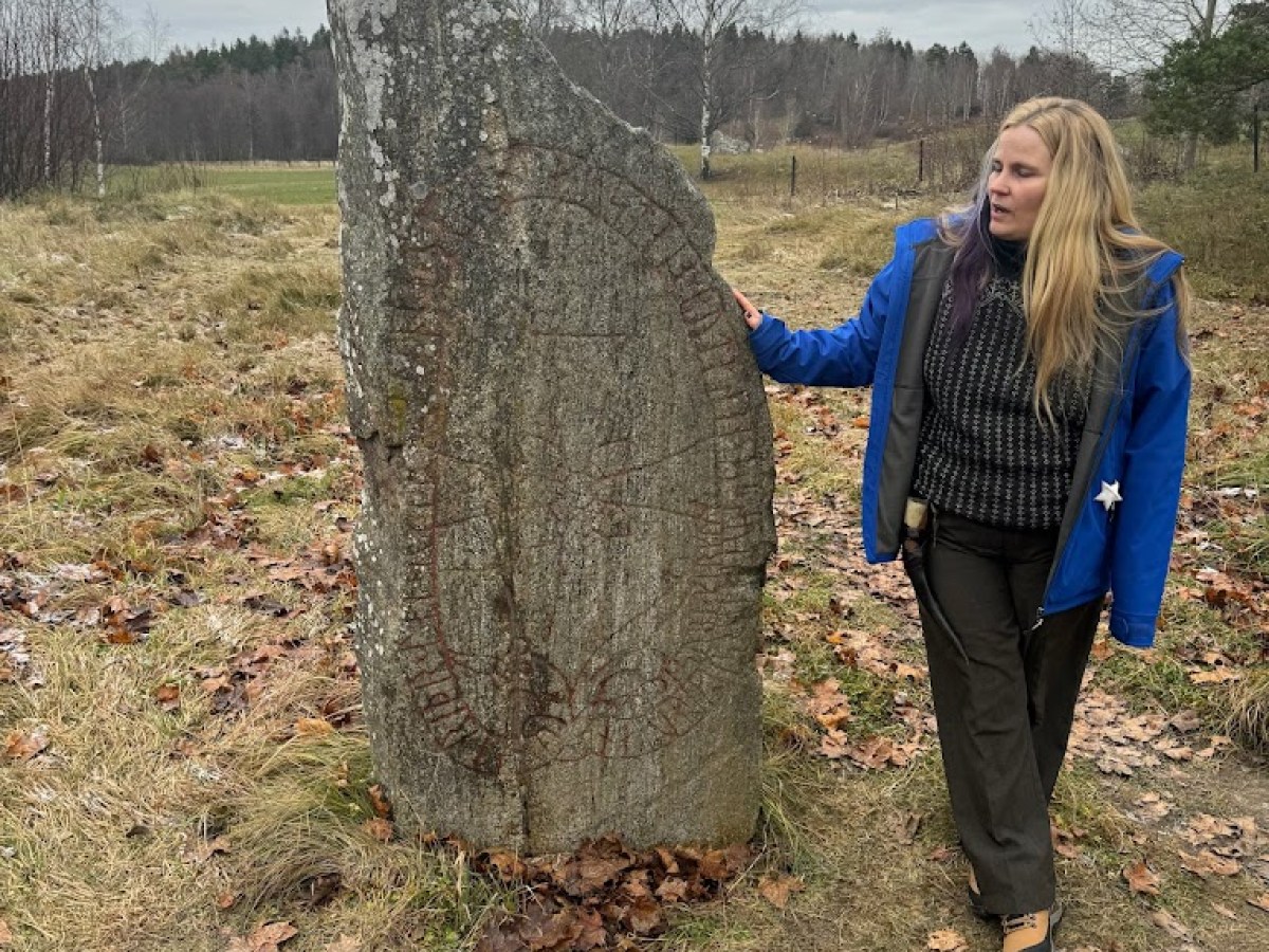 Person standing beside a tall, weathered rune stone in a grassy field.