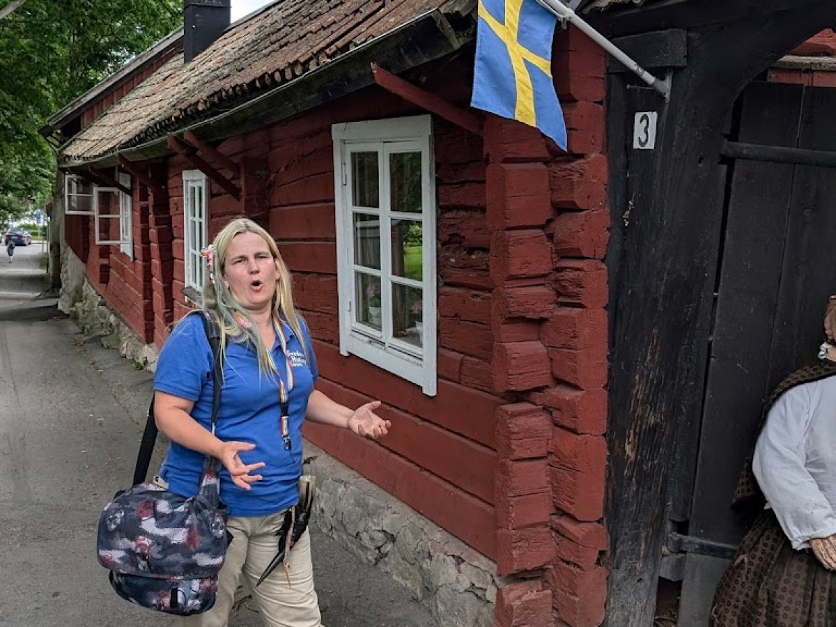 Woman in blue shirt gestures near rustic red house with Swedish flag.
