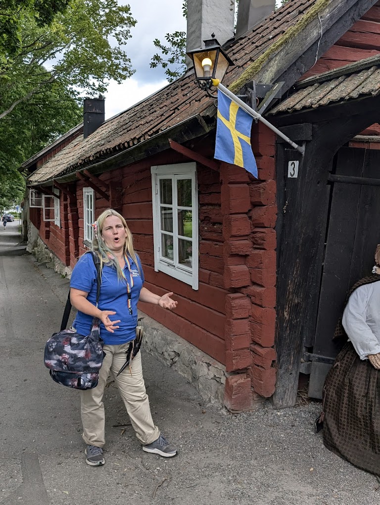 Woman in blue shirt gestures near rustic red house with Swedish flag.