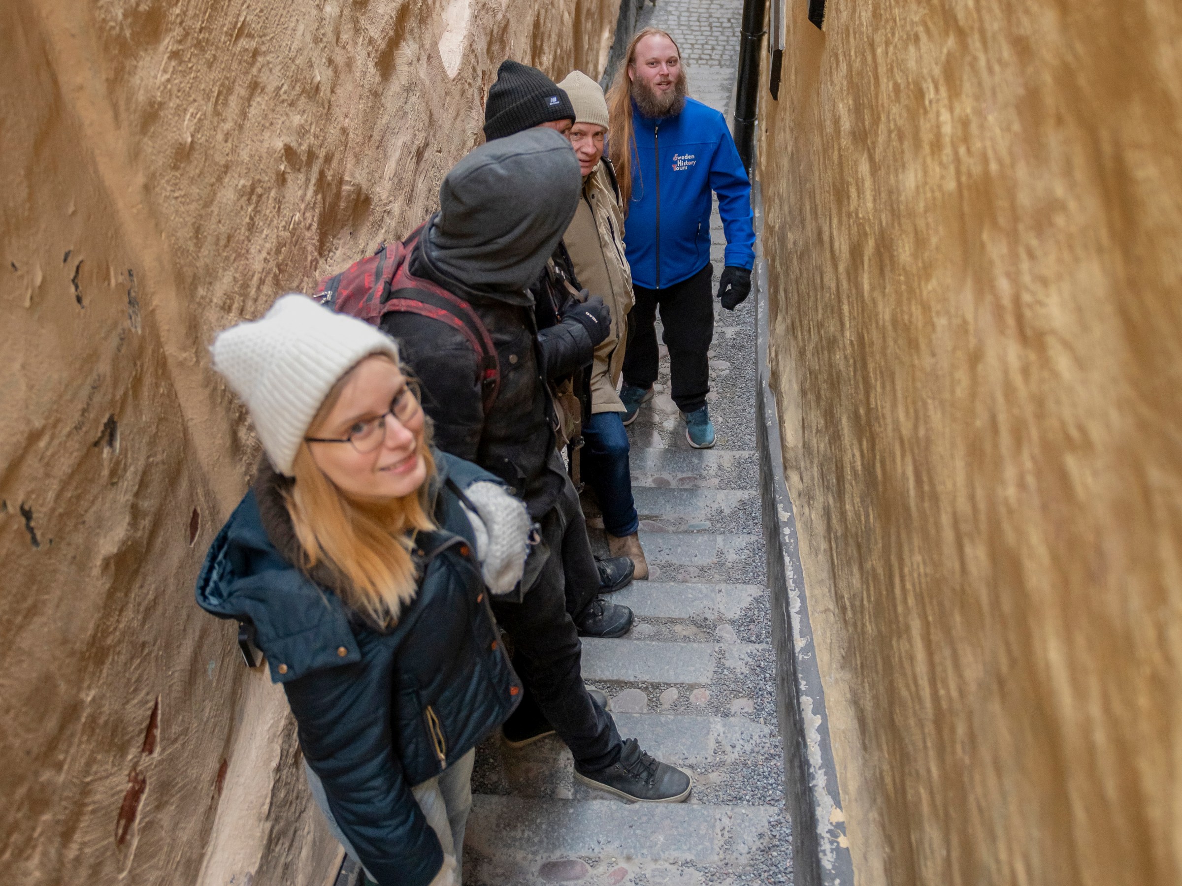 Four people in winter clothing walk down a narrow, stone stairway between tall walls.