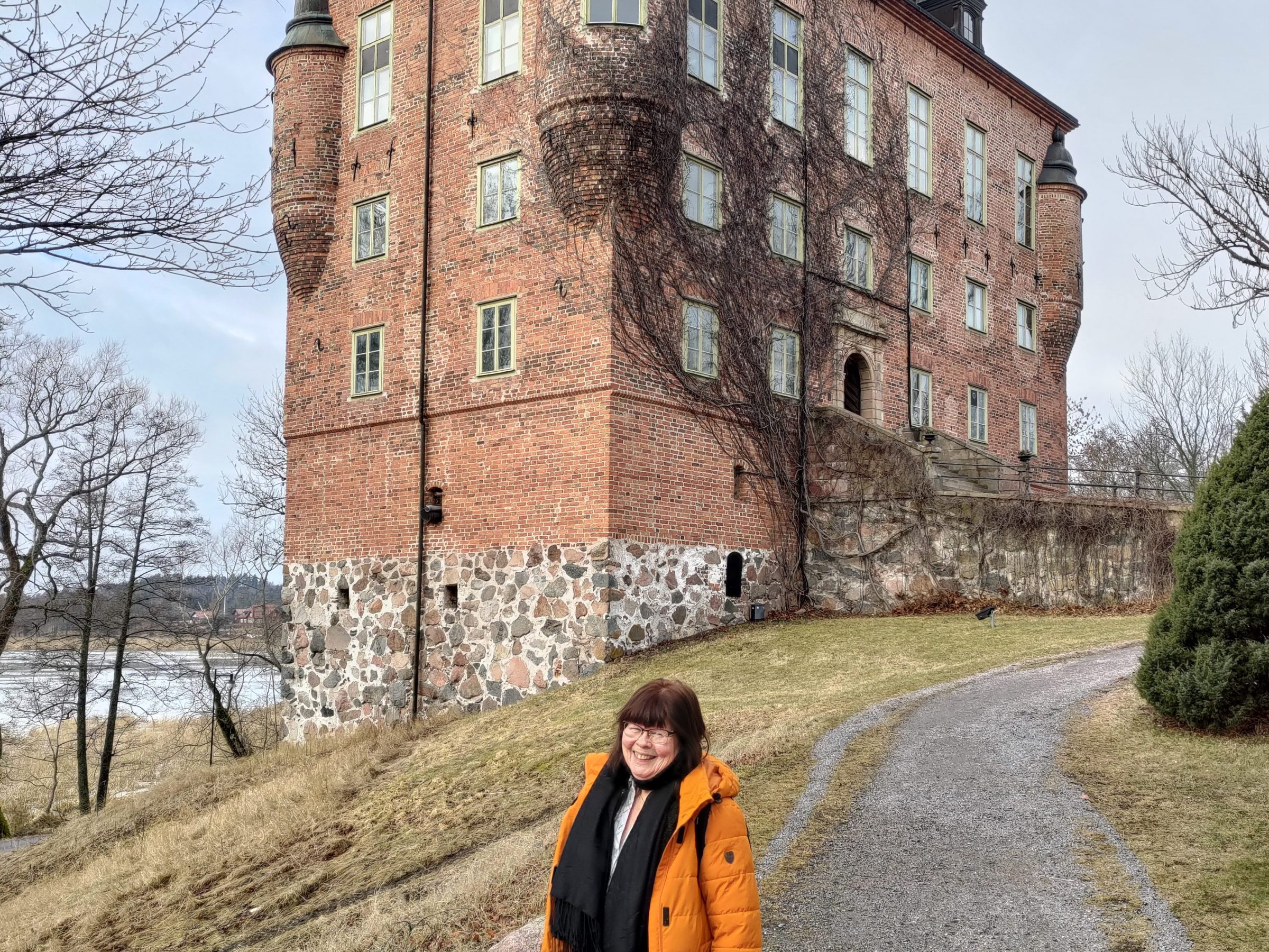 Woman in orange coat stands on path near a large, historic brick building.