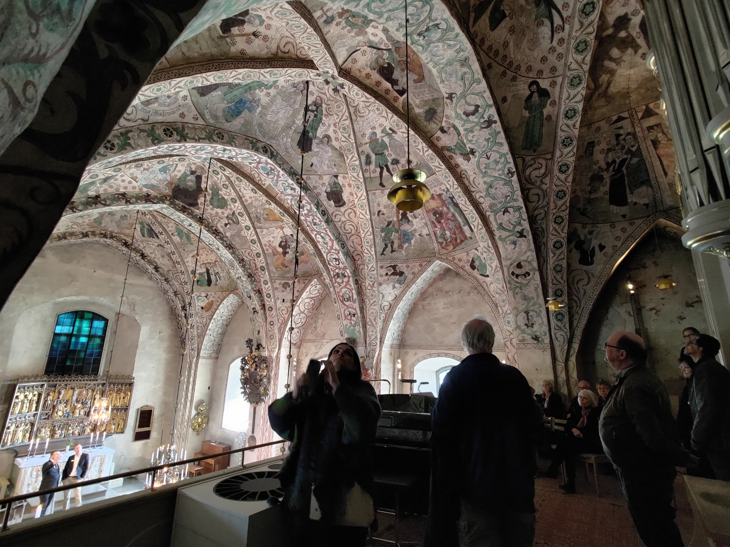 People inside a church with ornate painted vaulted ceiling and chandeliers.