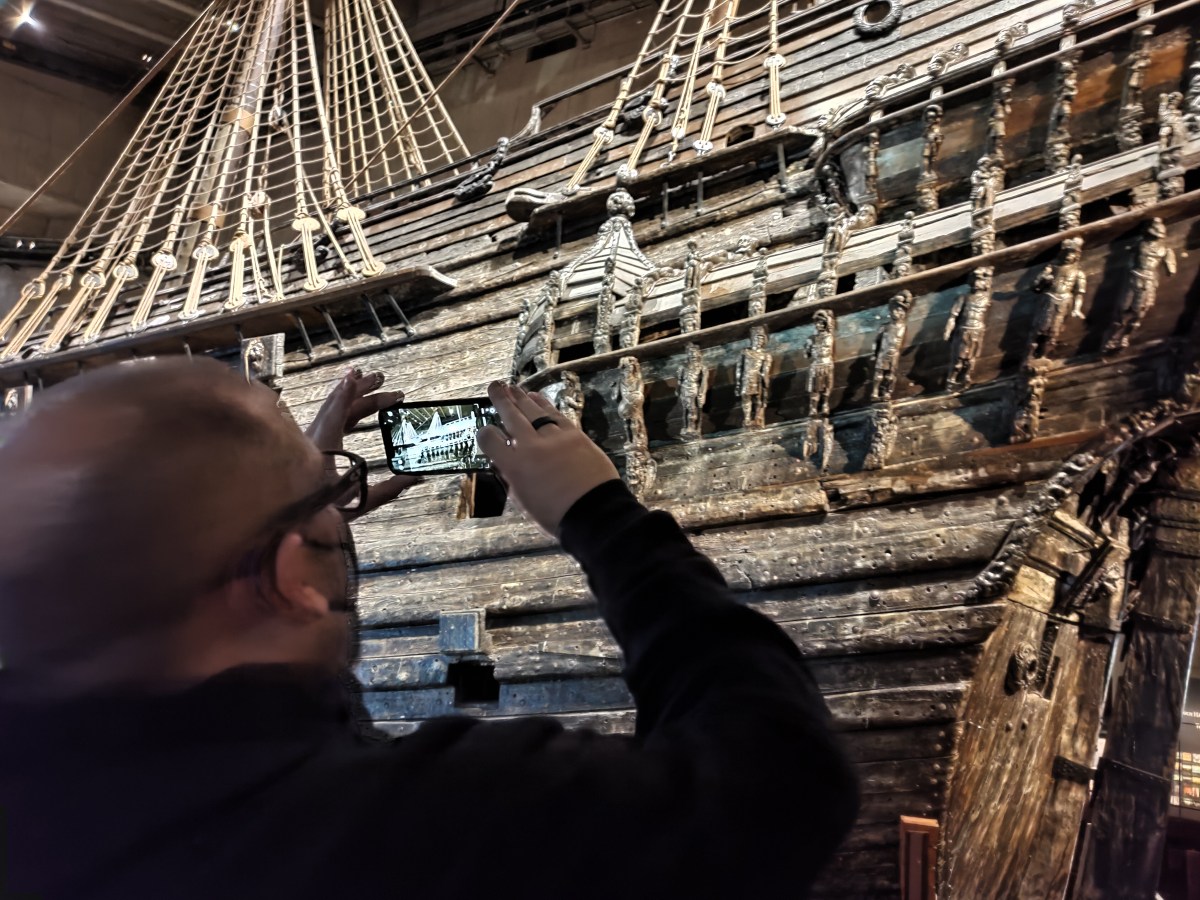 Person photographing a historic wooden ship with intricate carvings visible.
