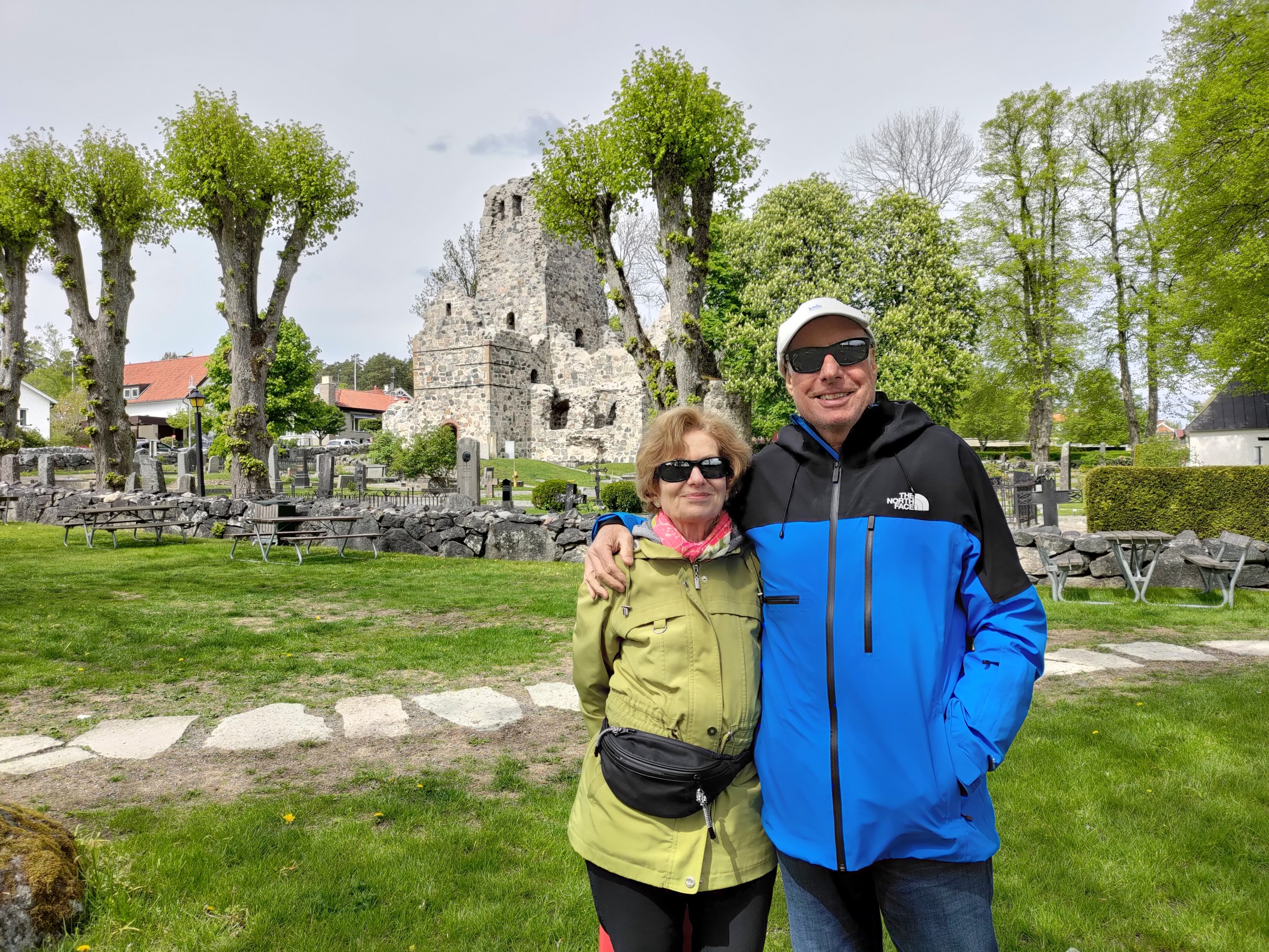 Two people in jackets smiling in front of ancient stone ruins and lush greenery.