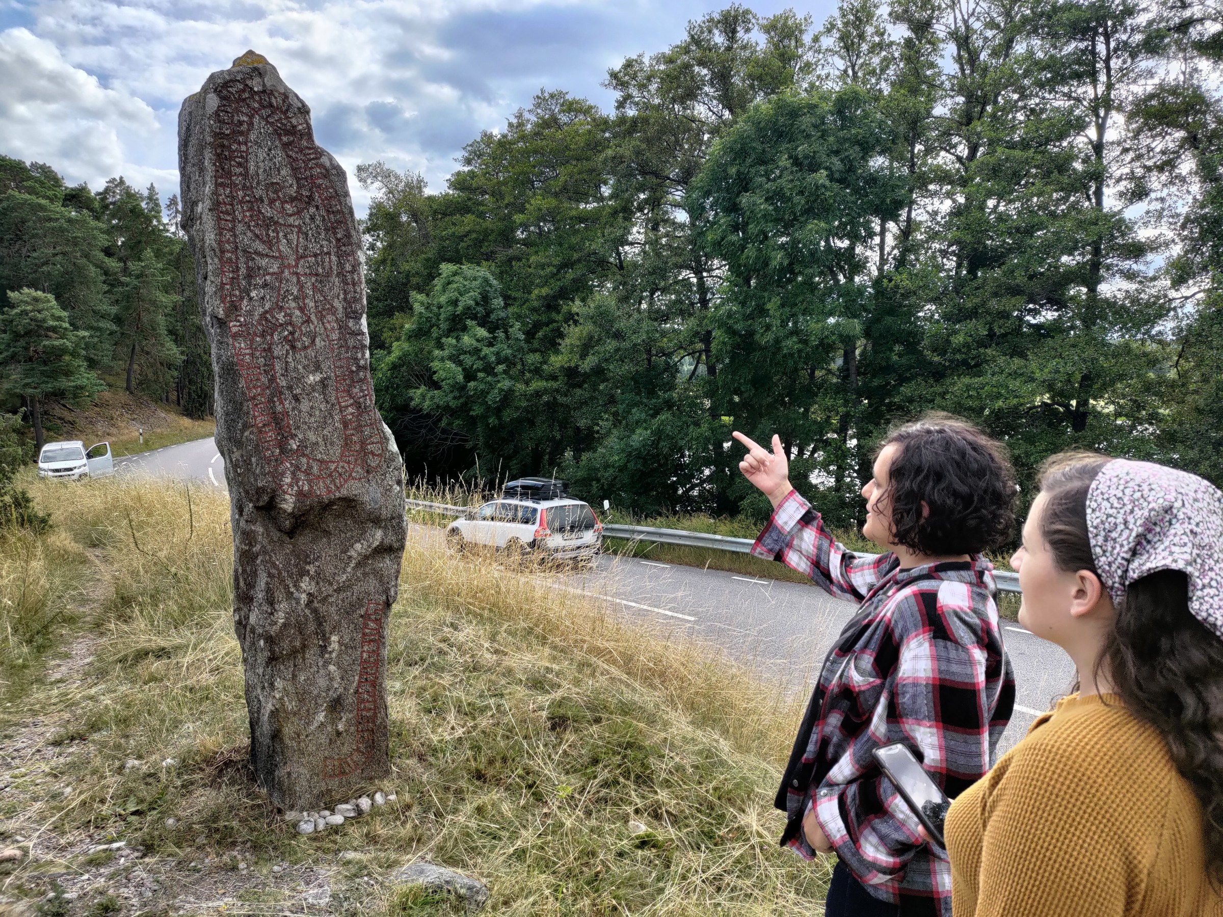 Two people observing a large runestone by a roadside, surrounded by trees and parked cars.