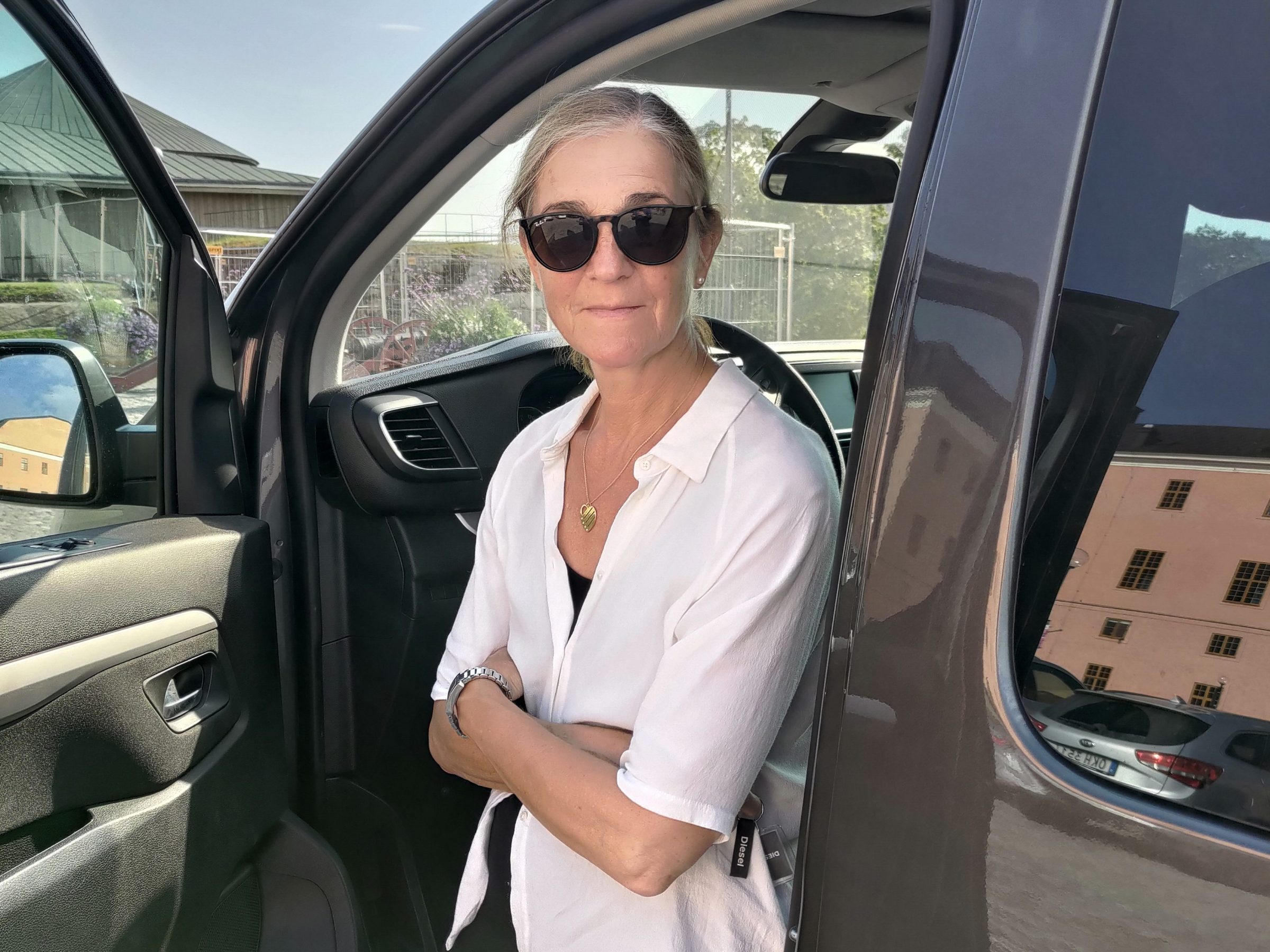 Woman with sunglasses in white shirt stands by an open car door on a sunny day.
