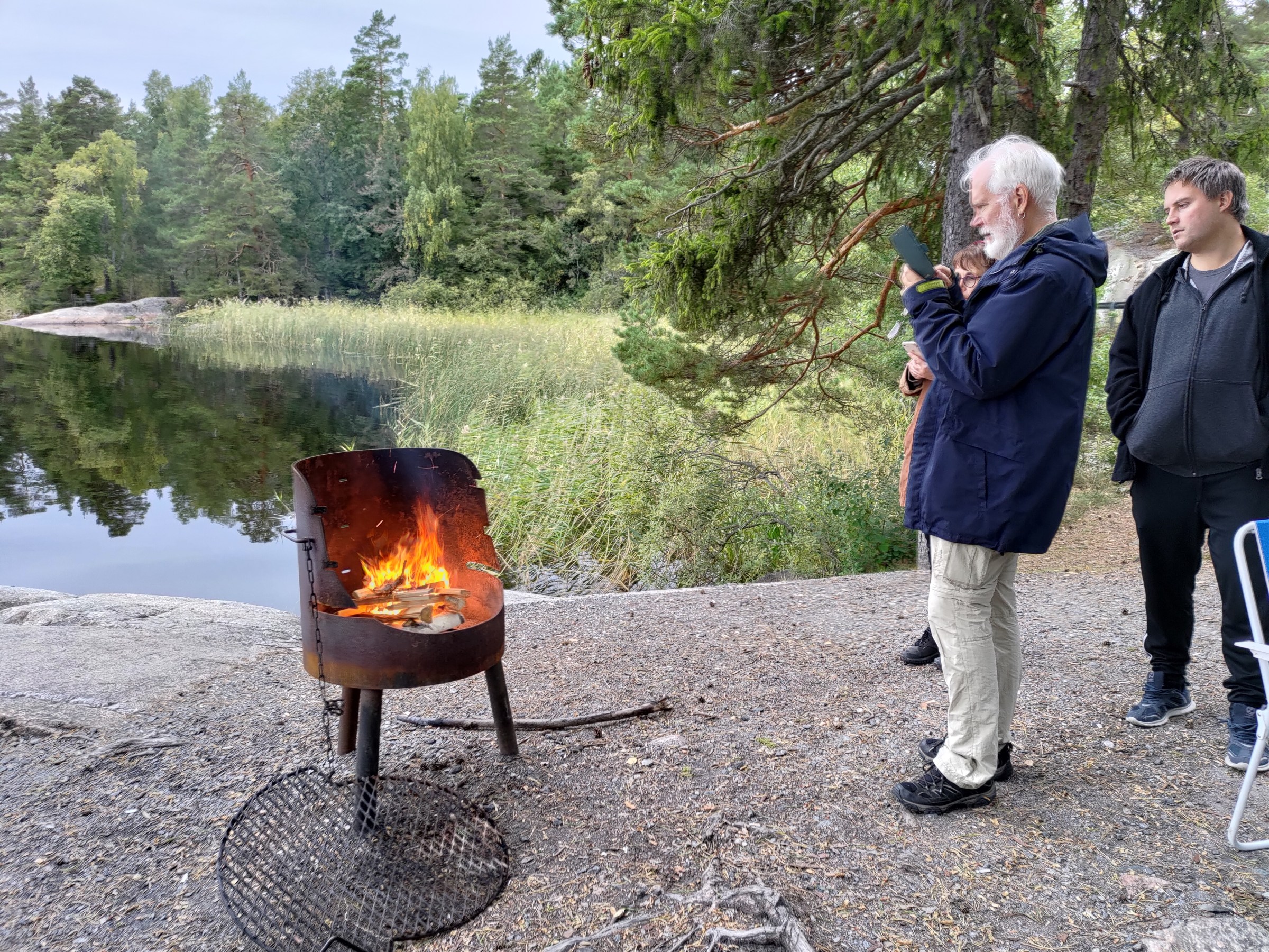 Two people by a lakeside with a metal fire pit burning on the ground.
