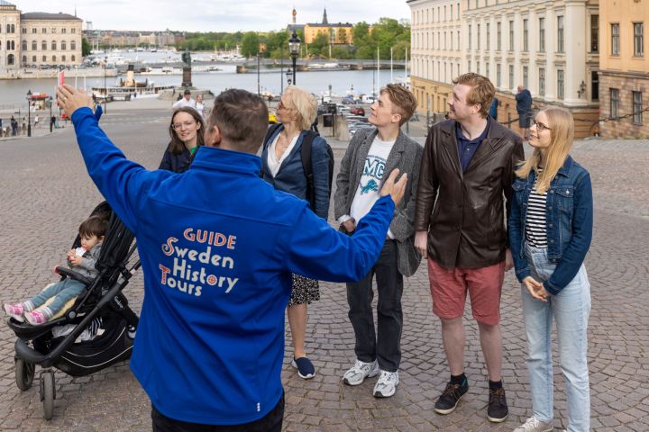 Tour guide speaks to a group in a historic city square with waterfront and buildings in the background.