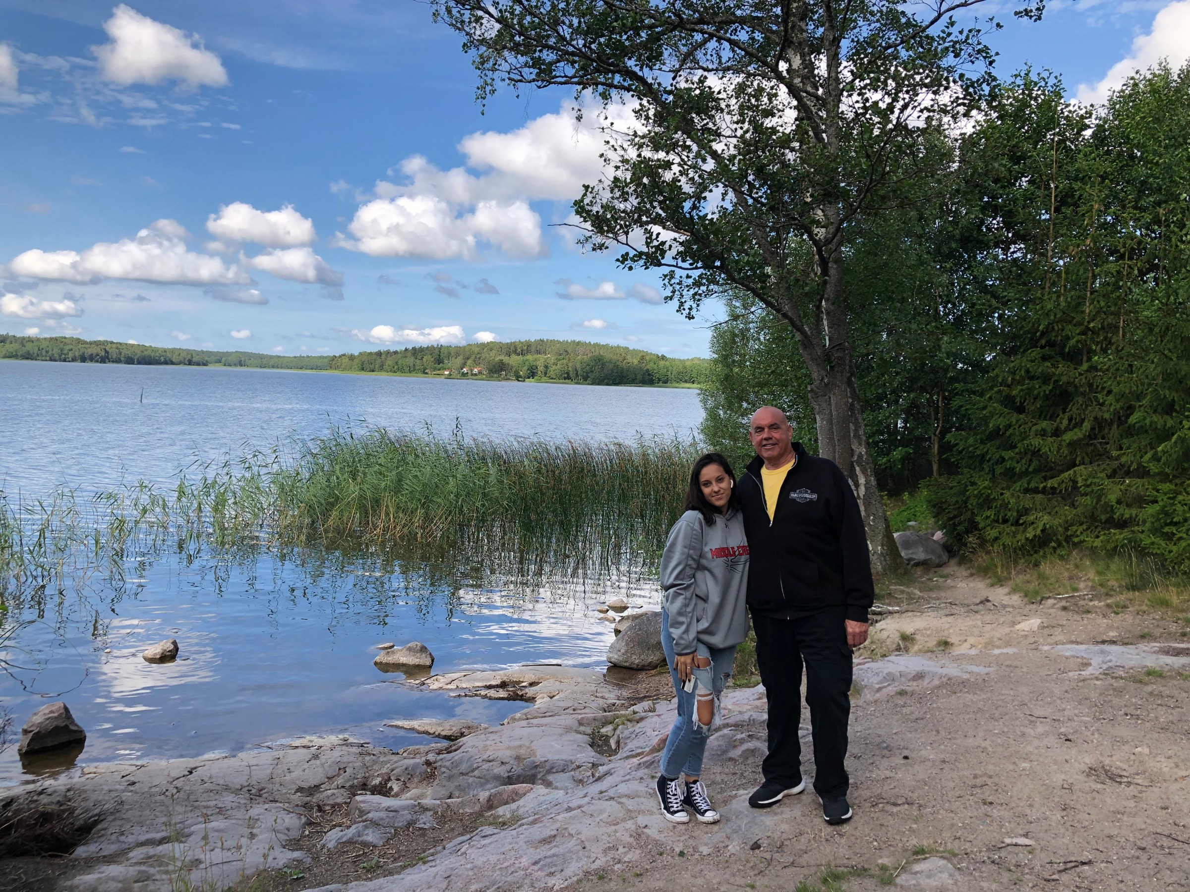 Two people standing by a lakeside with trees and clouds in the background.