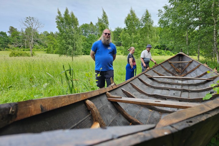 Three people stand near an old wooden boat in a grassy field with trees.