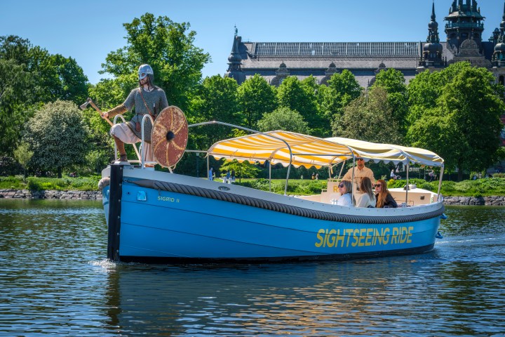 Blue sightseeing boat with passengers on a river, trees and building in background.